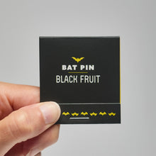 Hand holding a black matchbox labeled 'Bat Pin Black Fruit' with yellow bat graphics on a gray background.
